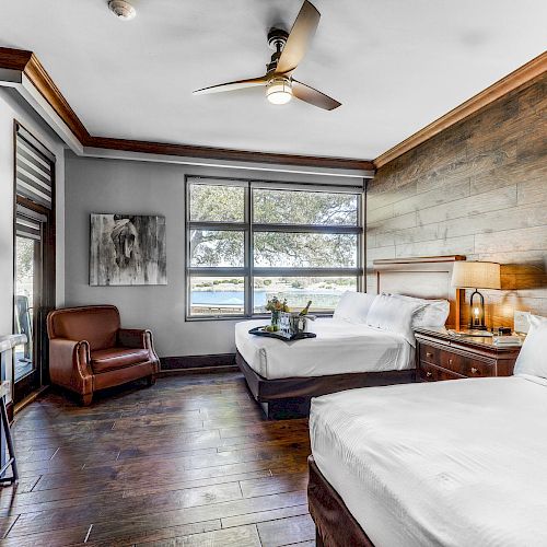 A hotel room features two beds, a wooden accent wall, a seating area, a desk, and a flat-screen TV. Large windows showcase an outdoor view.
