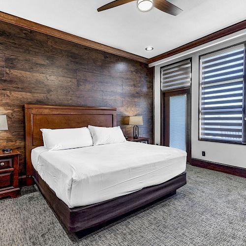 A modern bedroom with a wooden accent wall, a bed, nightstands with lamps, a ceiling fan, and large windows with blinds, ending the sentence.