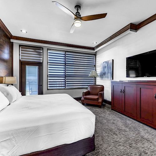 A modern bedroom features a large bed, TV, armchair, and a ceiling fan, with large windows showcasing a view.