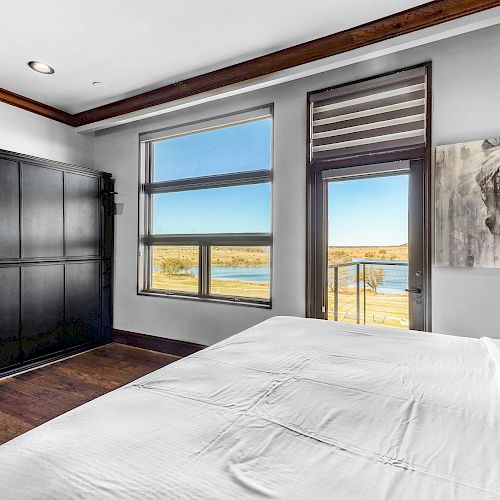 A bedroom with a neatly made bed, large windows showing a scenic view, a black wardrobe, and a horse painting on the wall.