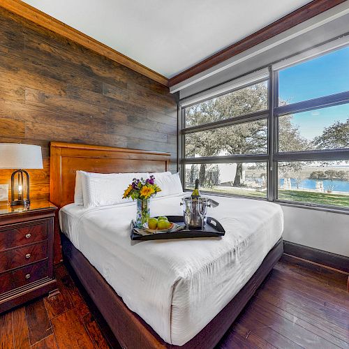A cozy bedroom with a wooden accent wall, a bed with a tray of flowers and a bottle, a nightstand with a lamp, and a window with a scenic view.