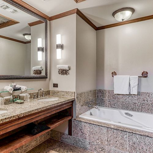 The image shows a bathroom with a double sink vanity, large mirror, wall lights, a jacuzzi tub, tiled walls, and a folded towel on a towel bar.