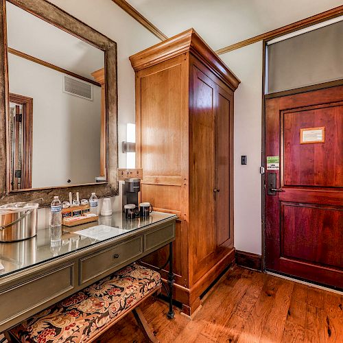 A hotel room entryway with wooden floors, a large mirror, a wardrobe, a wooden door, a desk with a mirror and several bottles of water.