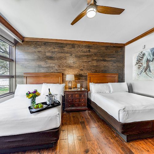 The image shows a cozy bedroom with two beds, a wooden accent wall, a nightstand, a ceiling fan, and a large window.