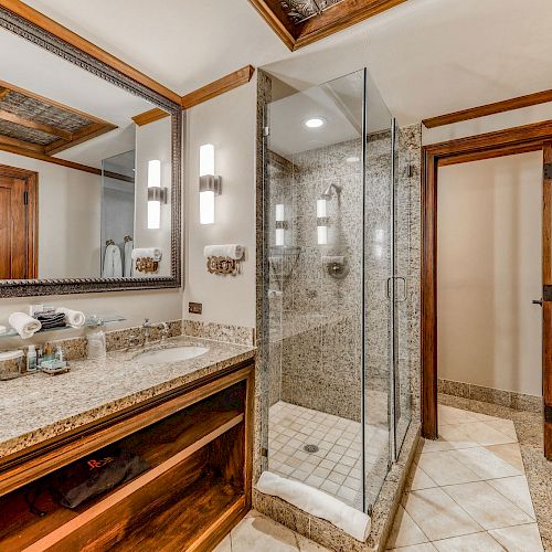 A modern bathroom with a granite countertop, double sinks, a glass-enclosed shower, and a wooden door. White towels hang on a wall rack.