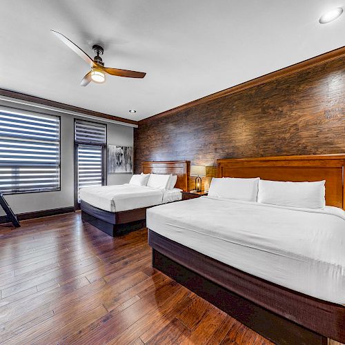 The image shows a modern bedroom with two large beds, wooden flooring, a ceiling fan, a bunk bed, and stylish dark wood furniture.