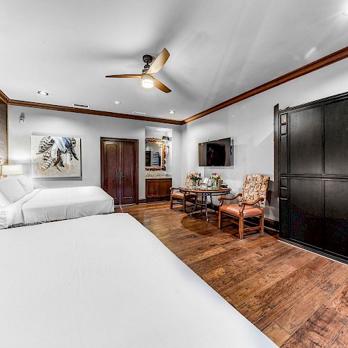 The image shows a hotel room with two beds, a ceiling fan, a TV, chairs, a table, and a painting on the wall, with wooden flooring ending the sentence.