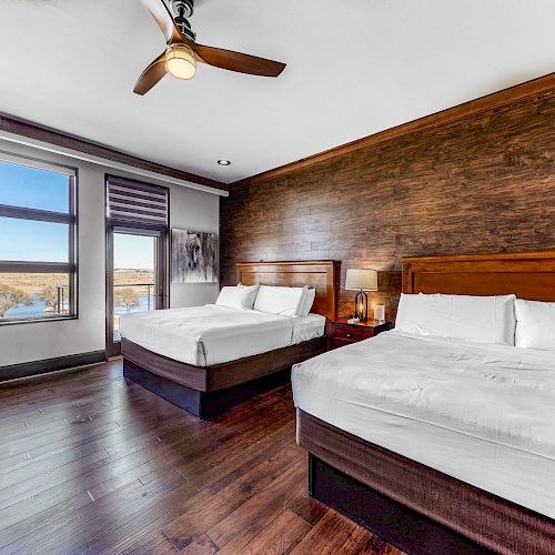 This image shows a modern hotel room with two large beds, wooden flooring, a ceiling fan, and a large window overlooking a scenic view.
