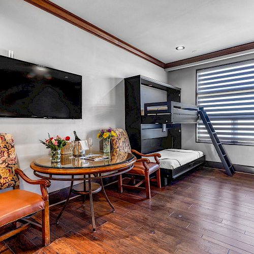 A room with a TV, chairs, a table with flowers and drinks, a bunk bed, and large window blinds, on wooden flooring.