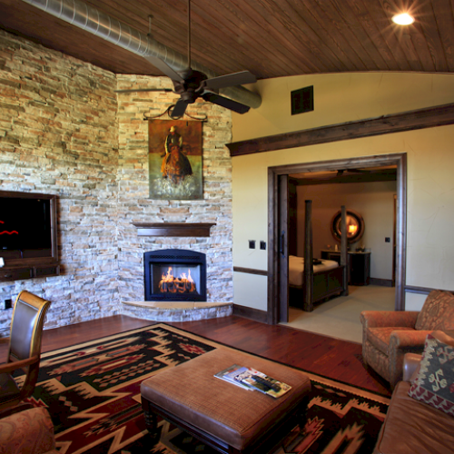 A cozy living room with a stone fireplace, flat-screen TV, rustic decor, wooden ceiling, and door opening to a bedroom with a bed and mirror.