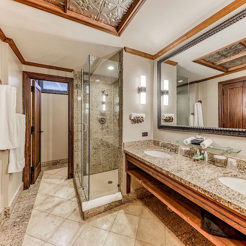 This image shows a luxurious bathroom with double sinks, a large mirror, a glass-enclosed shower, and elegant tile and wood details, ending the sentence.
