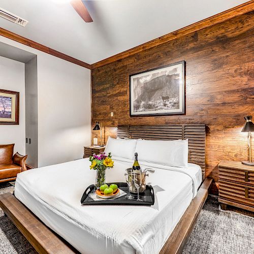A cozy bedroom with a wooden accent wall, a double bed with white linens, a tray with wine and fruits, a leather chair, and framed artwork.