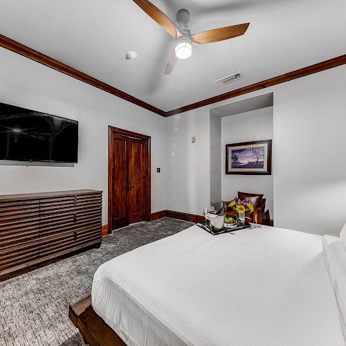 The image shows a modern bedroom with a bed, a TV on the wall, a dresser, a ceiling fan, and a tray with items on the bed.