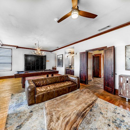 A spacious living room features brown leather sofas, wooden furniture, a ceiling fan, a pool table, and a horse painting on the wall.
