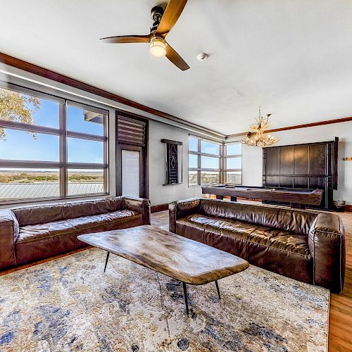 A spacious living room with large windows, two brown leather sofas, a wooden coffee table, a ceiling fan, and a chandelier, offering a scenic view.
