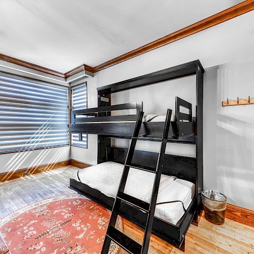 The image shows a room with a black bunk bed, large windows with blinds, a wooden bench, a red patterned rug, and wooden elements in the decor.