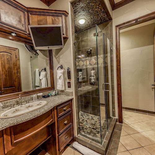 A modern bathroom featuring a granite countertop, wooden cabinets, a mounted TV, a glass-enclosed shower, and a towel rack with a towel.