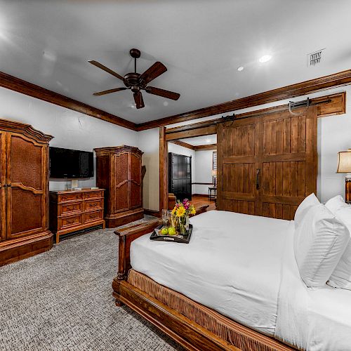 A cozy bedroom with wooden furniture, a bed, nightstands, a dresser, a TV, a ceiling fan, and a tray with flowers on the bed.