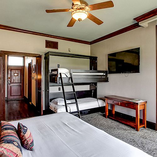 A cozy hotel room with a bunk bed, a double bed, a wall-mounted TV, a ceiling fan, an open bathroom door, and a balcony door.