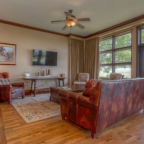 A cozy living room with leather sofas, a TV mounted on the wall, large windows, a ceiling fan, and wooden floors, exuding a rustic charm.