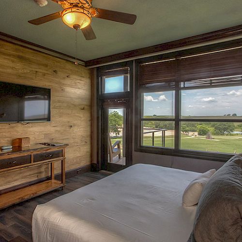 A cozy bedroom with a bed, TV, desk, ceiling fan, and large window offering a scenic view of the outdoors.