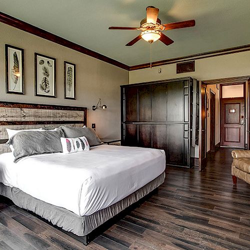 A modern bedroom with a king-sized bed, ceiling fan, wooden decor, wall art, a leather chair, side tables, and a wardrobe. The sentence ends.