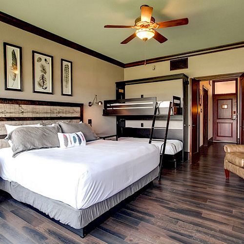 A well-furnished hotel room with a double bed, bunk beds, a ceiling fan, and a sitting area, featuring modern decor and wooden flooring.