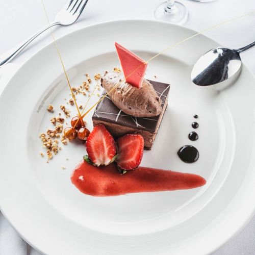 A chocolate dessert with strawberries, sauce, and a decorative element on a white plate, accompanied by a fork and spoon.