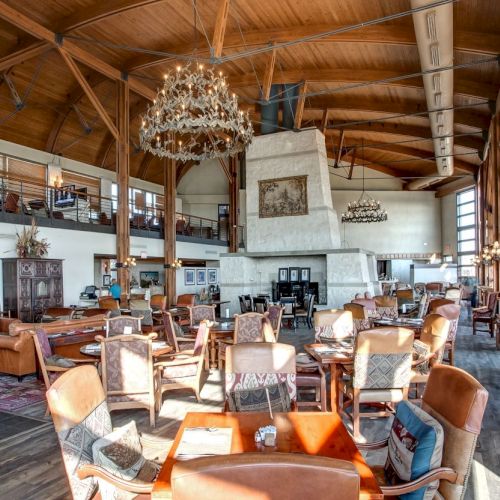 The image shows a spacious, rustic restaurant with wooden beams, chandeliers, and a large stone fireplace, filled with tables and chairs.