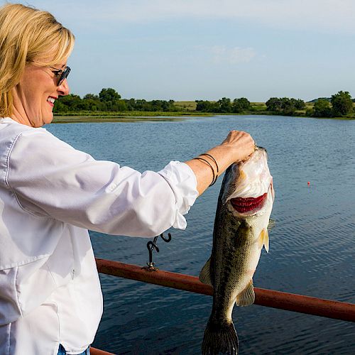 A person stands near a body of water, holding a large fish with a smile on their face, enjoying the moment outdoors.