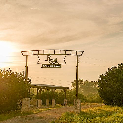 The image shows a pathway leading to an entrance marked by a sign that reads "R&R Sporting Clays," surrounded by greenery under a sunset sky.