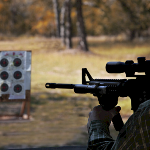 A person using a scoped rifle is aiming at a target board with multiple bullseyes in an outdoor setting with trees in the background.