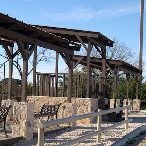 The image shows an outdoor seating area with a wooden pergola, benches, stone walls, and overhead lights near a gravel path.