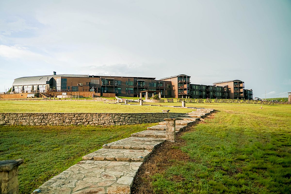 The image shows a modern building complex with a stone pathway leading up to it, set against a grassy landscape under a cloudy sky.