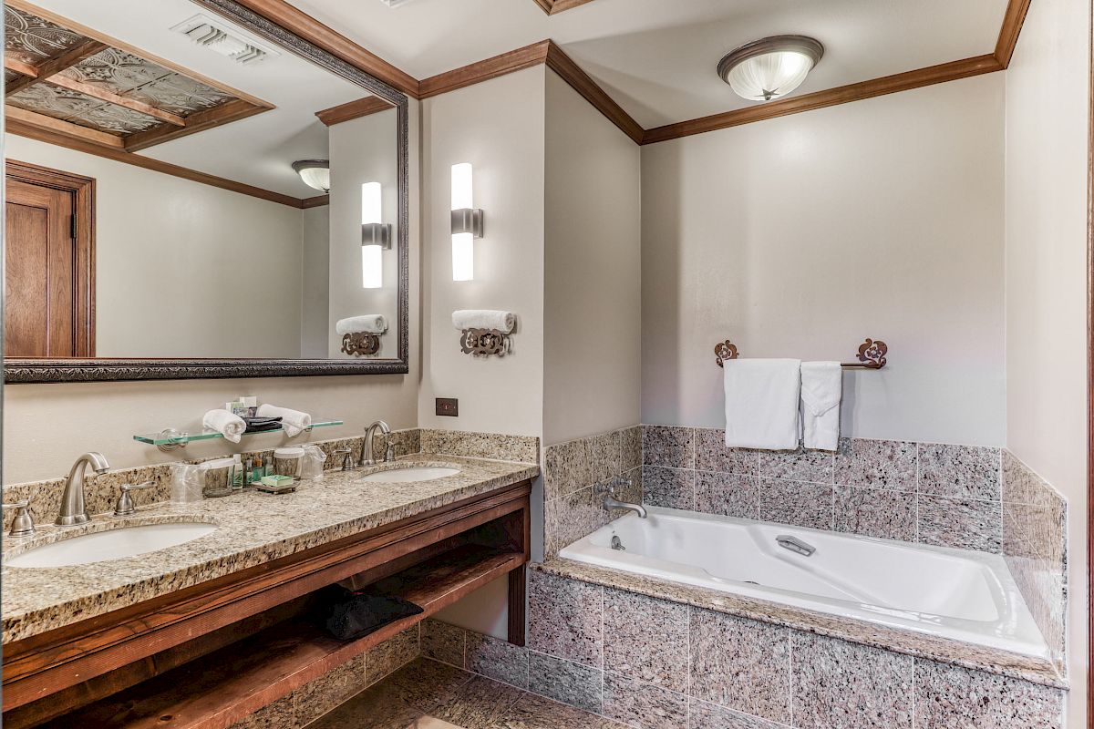 This image shows a bathroom with a double sink vanity, mirrors, lighting fixtures, and a bathtub with a towel hanging above it on the wall.