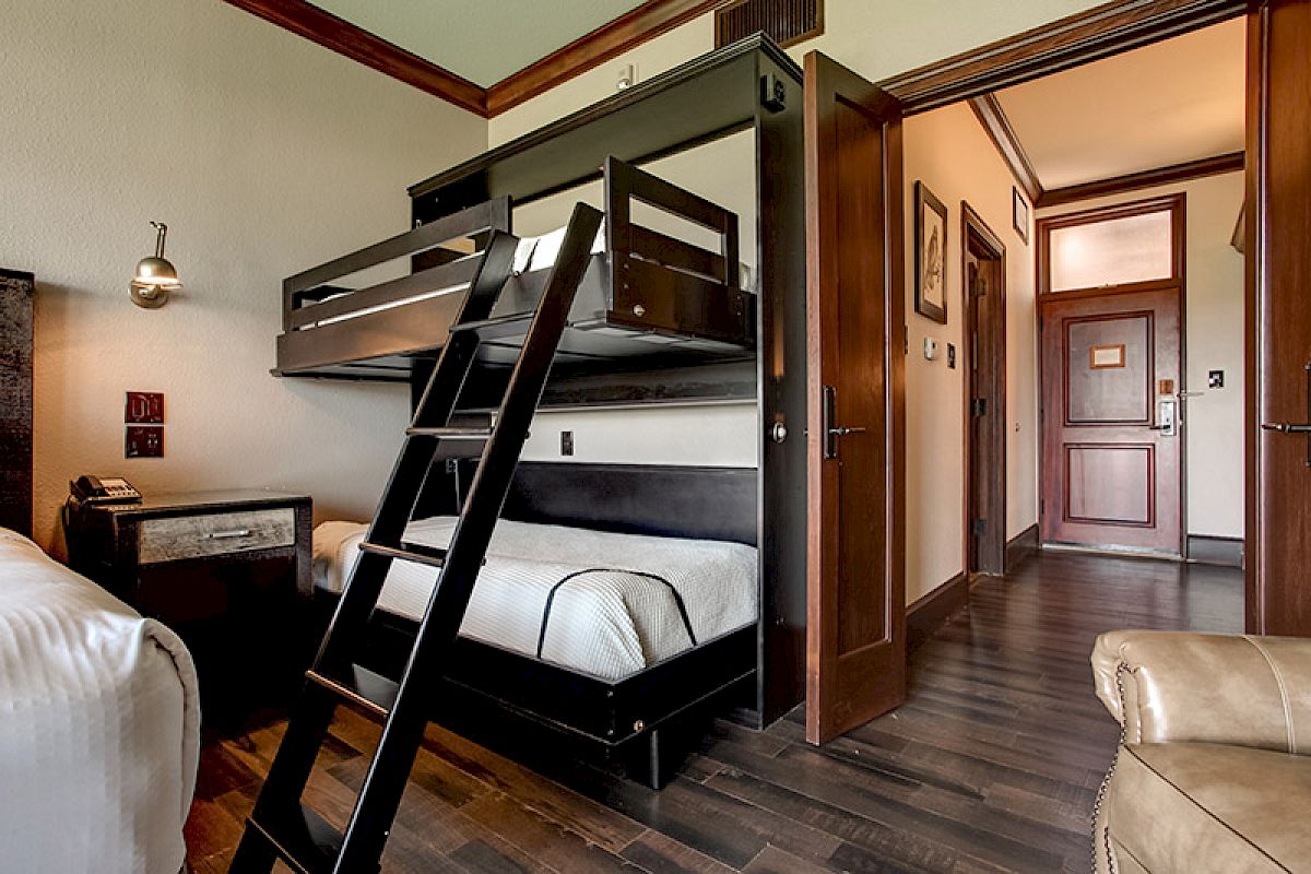 The image shows a cozy bedroom with bunk beds, wooden flooring, and a small desk with a wall lamp. The room has an open-door view of a hallway.