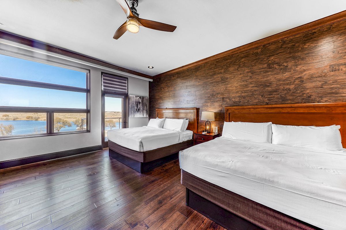 A hotel room with two large beds, wooden flooring, a window with a scenic view, and a ceiling fan.