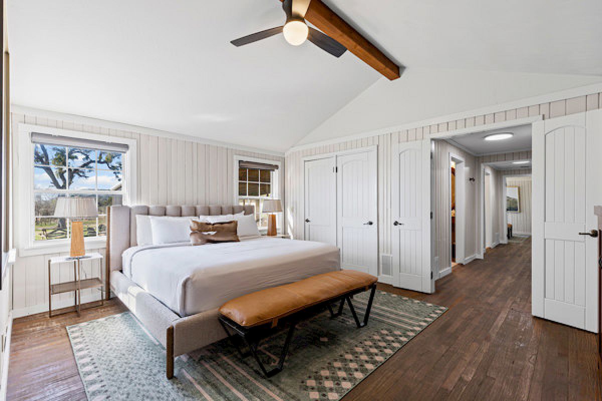 A cozy bedroom with a large bed, wooden floor, ceiling fan, and bright windows offering a scenic view and ample natural light.