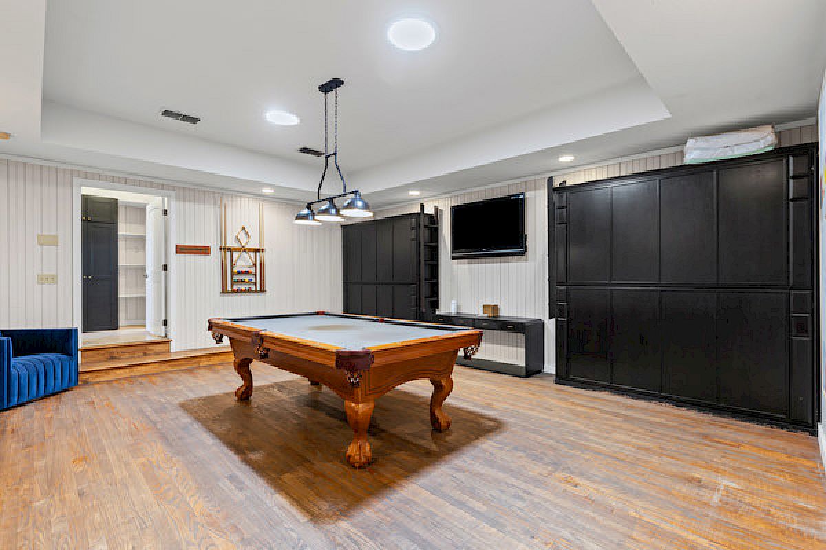 A game room with a pool table, wooden floor, wall-mounted TV, closed door, cue rack, and ceiling lights.