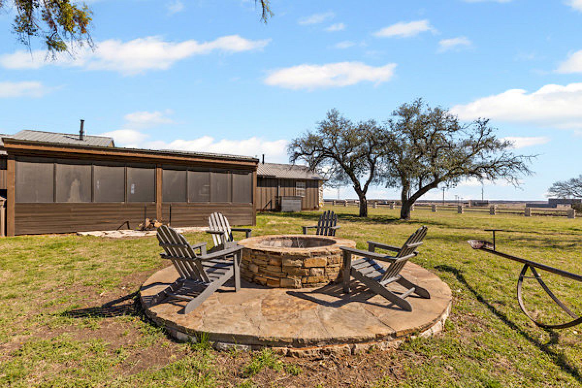 A rustic outdoor scene with a circular fire pit, surrounded by chairs, near a cabin and trees under a clear sky.