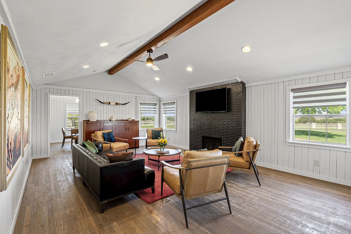 A stylish living room with a black brick fireplace, wall-mounted TV, sofas, and wooden beams on the ceiling, featuring large windows.