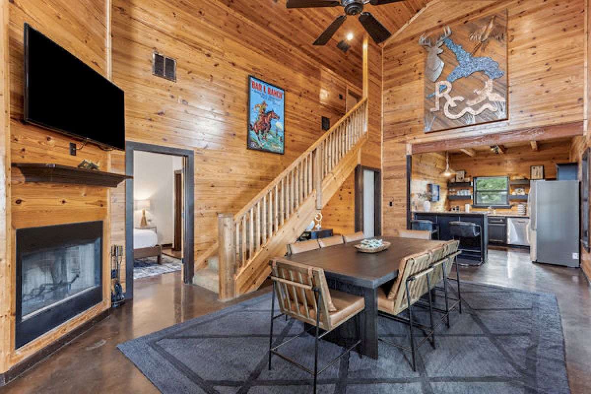 A cozy cabin interior with wooden walls, dining table, chairs, mounted TV, fireplace, staircase, and kitchen. Rustic decor throughout.