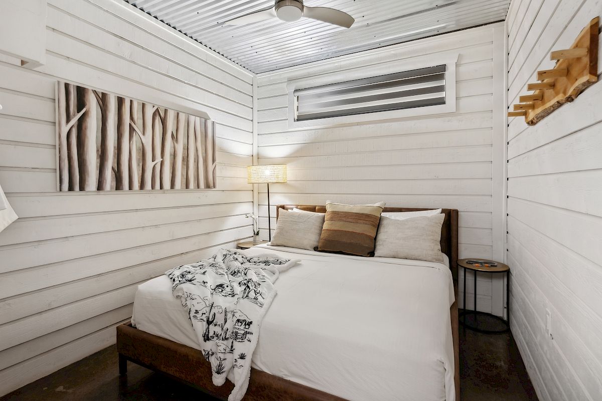 A cozy bedroom with wood-paneled walls, a bed with cushions, a blanket, side table, artwork, and a ceiling fan.