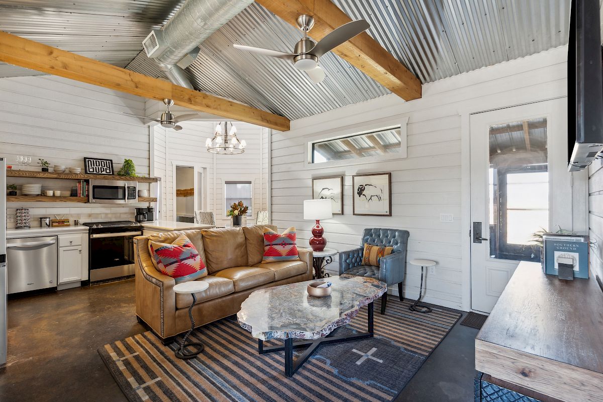 A cozy living room with wood beams, a leather sofa, a marble coffee table, and rustic decor. Kitchen area visible in the background.