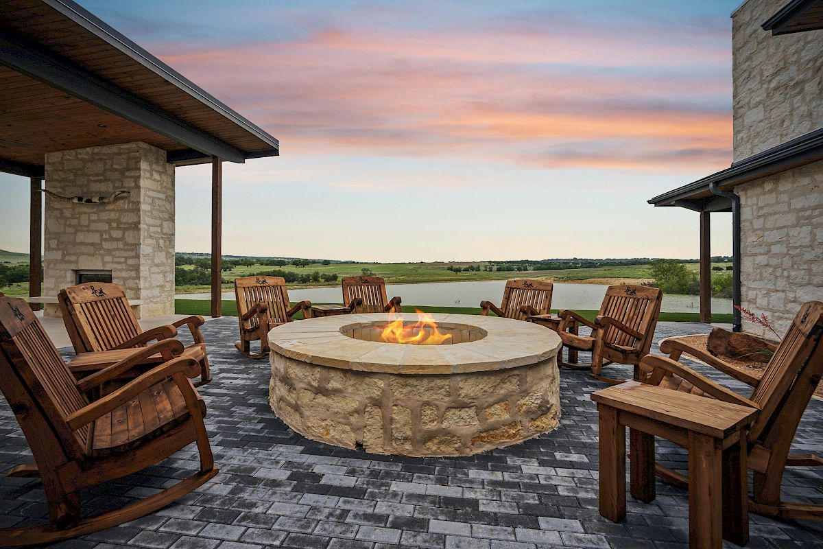 Outdoor patio with a central fire pit, surrounded by wooden chairs, stone walls, and scenic countryside view at sunset.