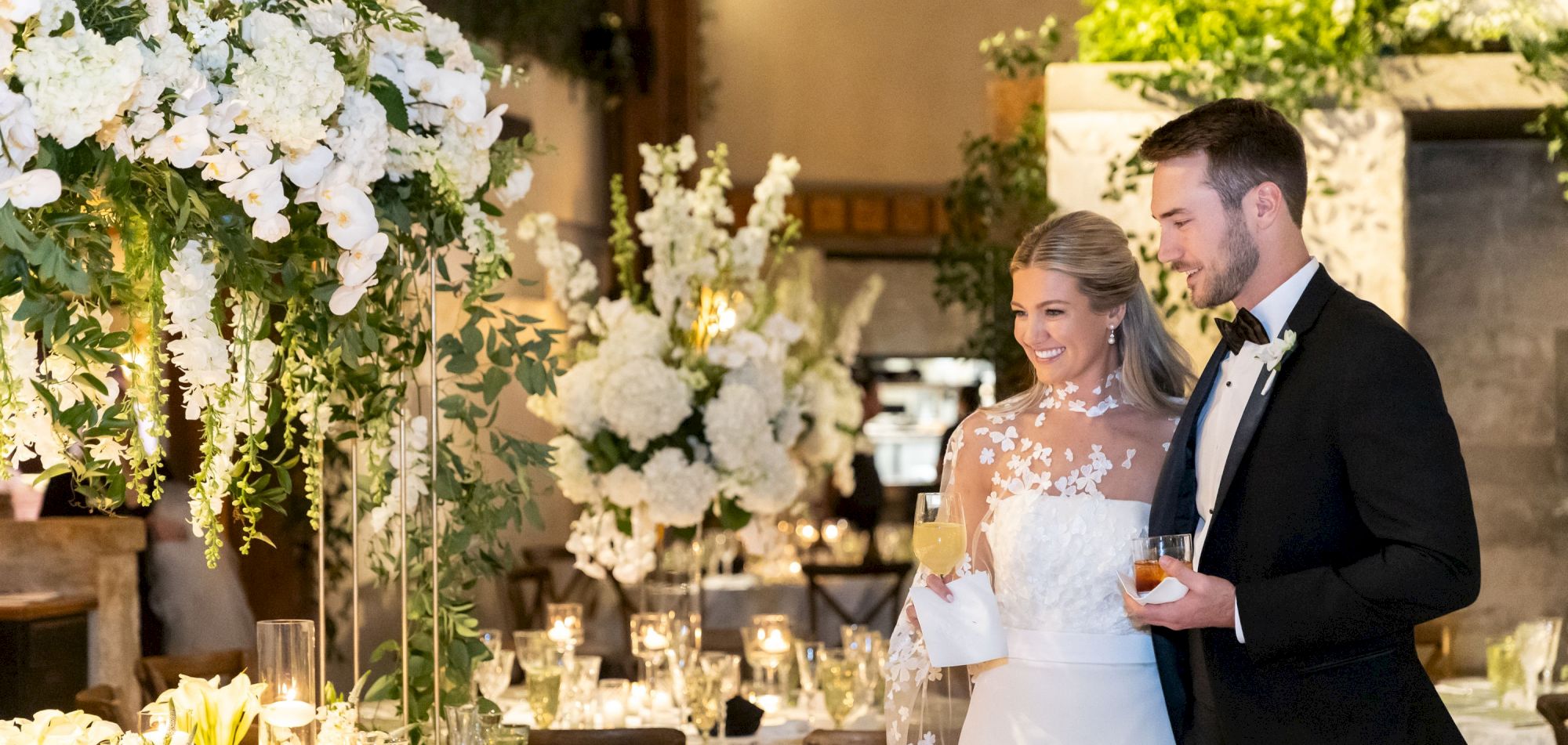 A couple at a wedding reception, surrounded by elegant floral arrangements and lit candles, with drinks in hand.