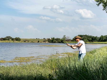 A man wearing a hat is fishing by a lake, surrounded by grass and trees under a partly cloudy sky.