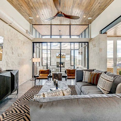 A modern living room with large windows, a comfortable seating area with gray couches, a TV, wooden ceiling, and a stylish fan.