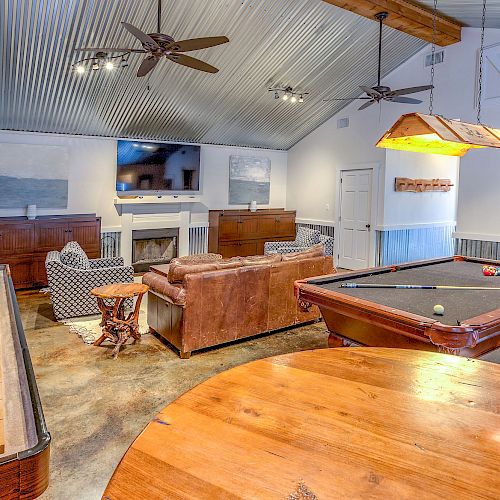 A cozy game room with a pool table, shuffleboard table, couch, chairs, a TV, ceiling fans, and various decorations on the walls.