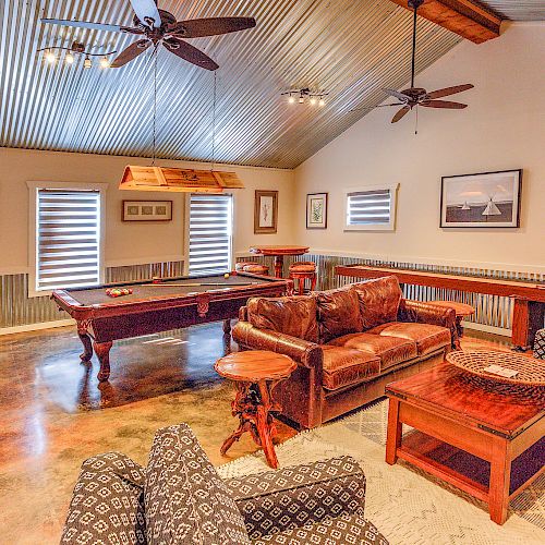 The image shows a cozy living room with a pool table, several sofas, a coffee table, ceiling fans, and framed art on the walls, creating a warm ambiance.